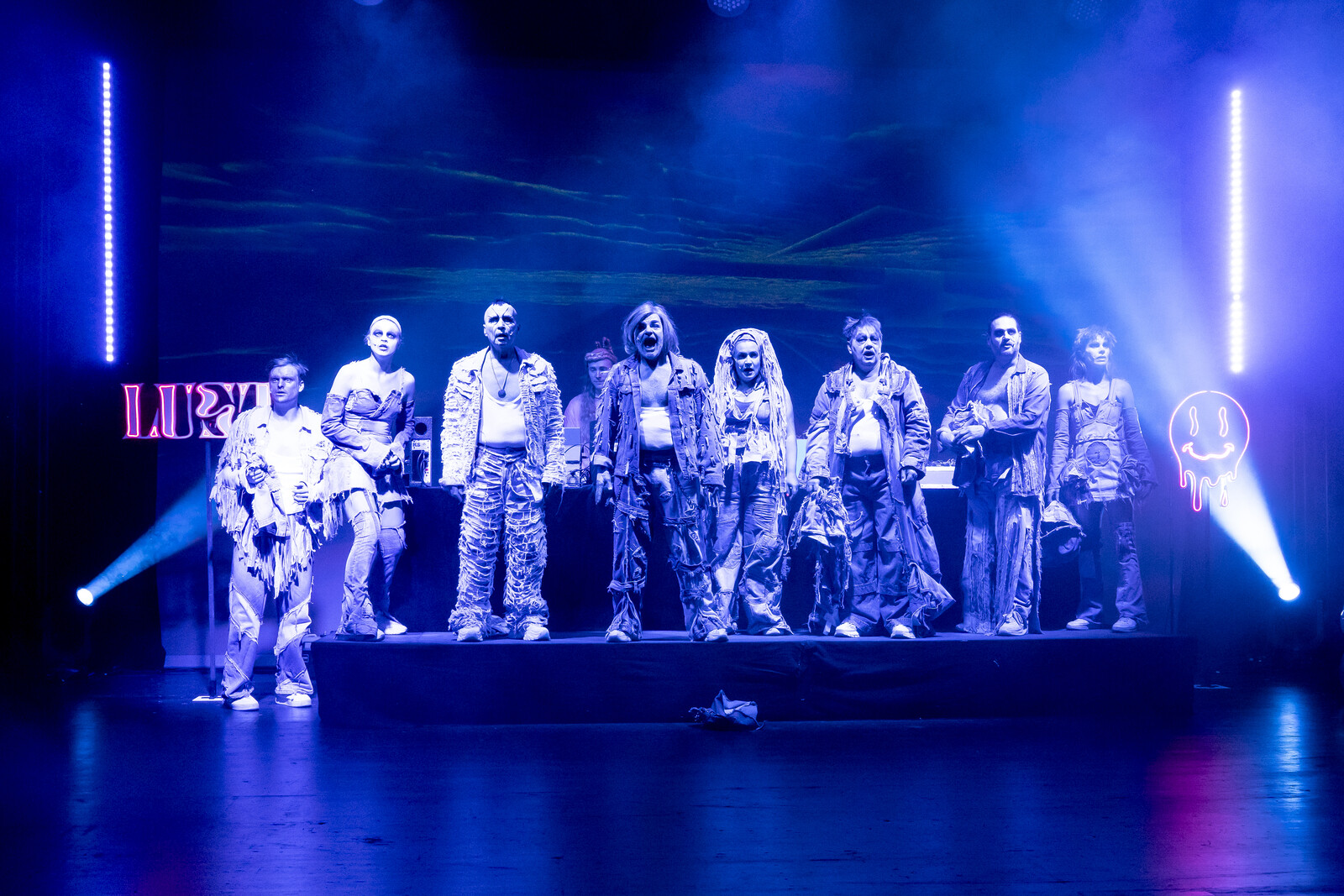 Die Gruppe Schauspieler*innen steht aufgereiht vor dem DJ-Pult in blauem Licht.
