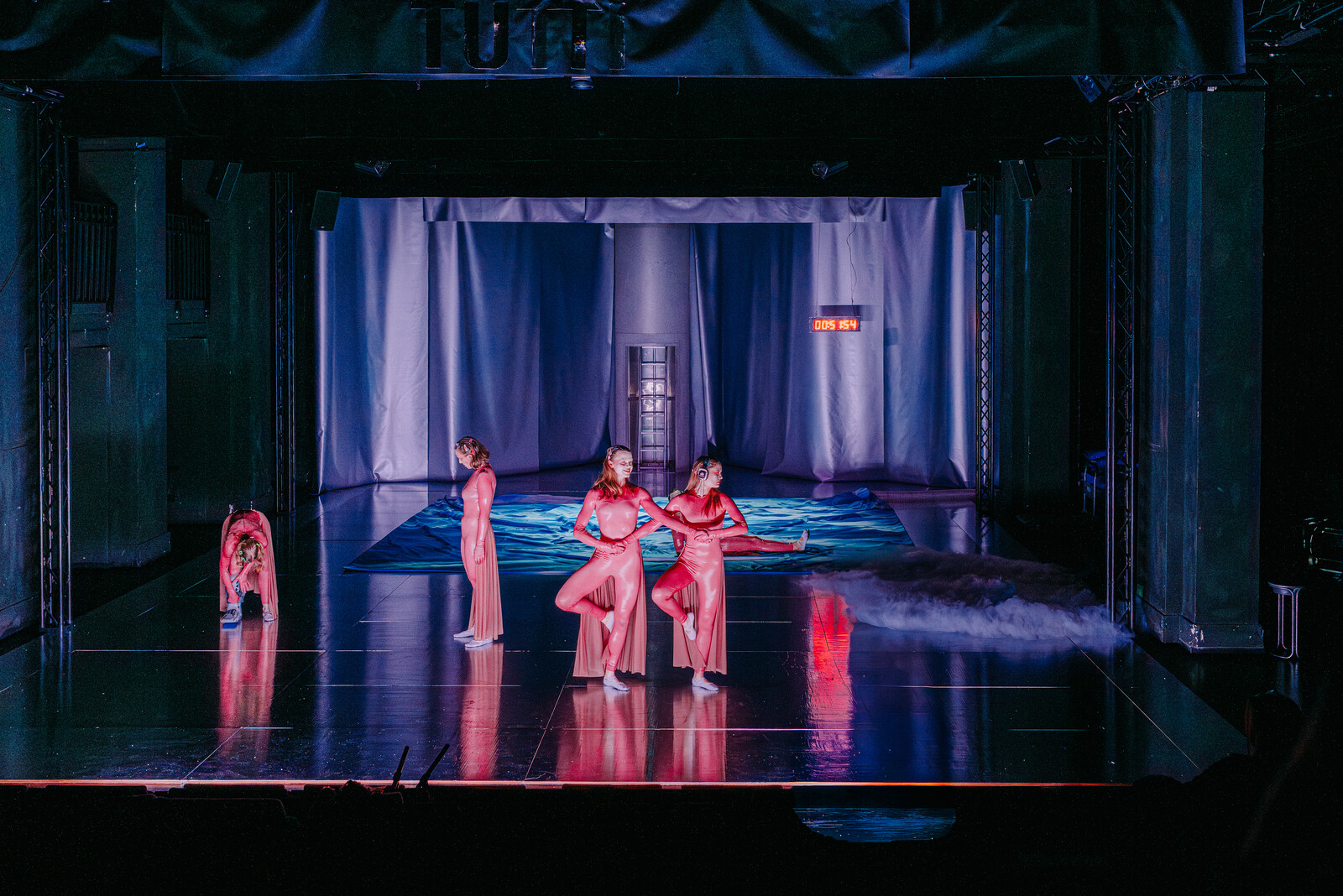 Bühnensituation im lila Licht mit vier Schauspieler:innen im Vordergrund in rosa Kostümen.