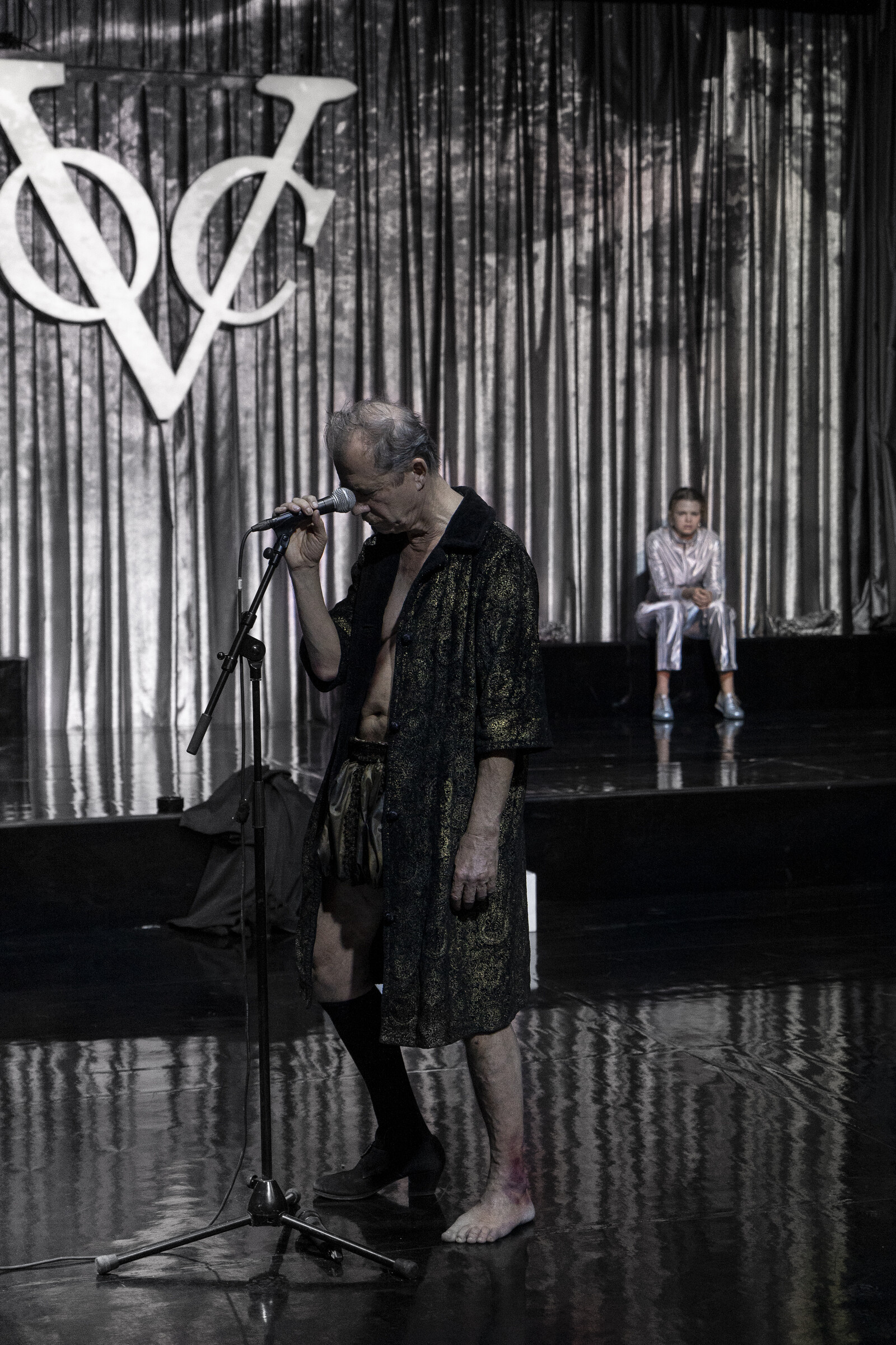 Judge Adam in a dressing gown stands in front of a microphone stand, behind him sits Eve in a silver jogging suit.