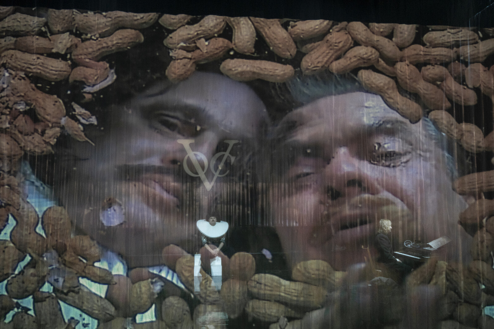Two men, projected onto a gauze, look through a pile of peanuts into the audience.