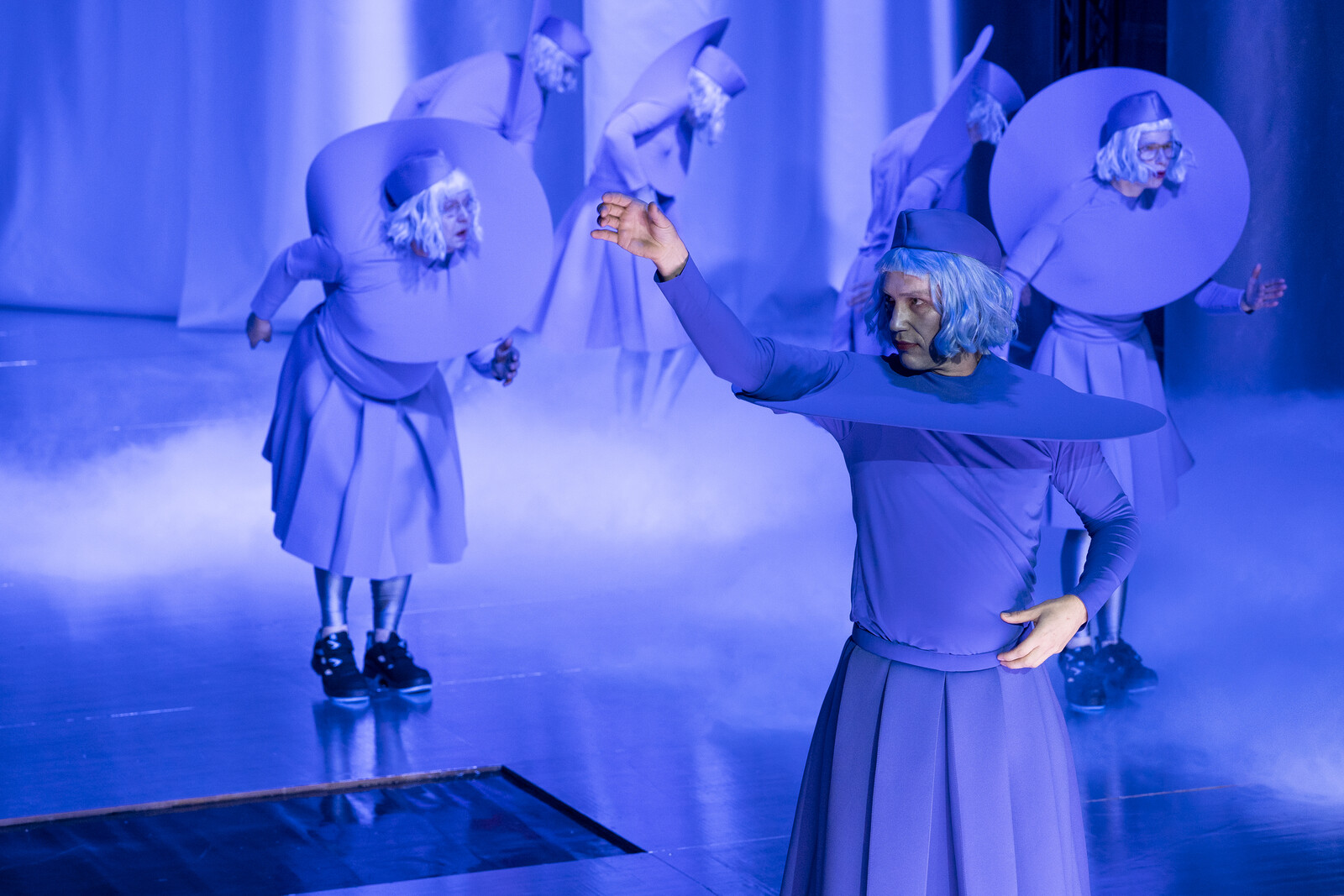 Bernhard Glose stands in the foreground while the ensemble dances in the background.