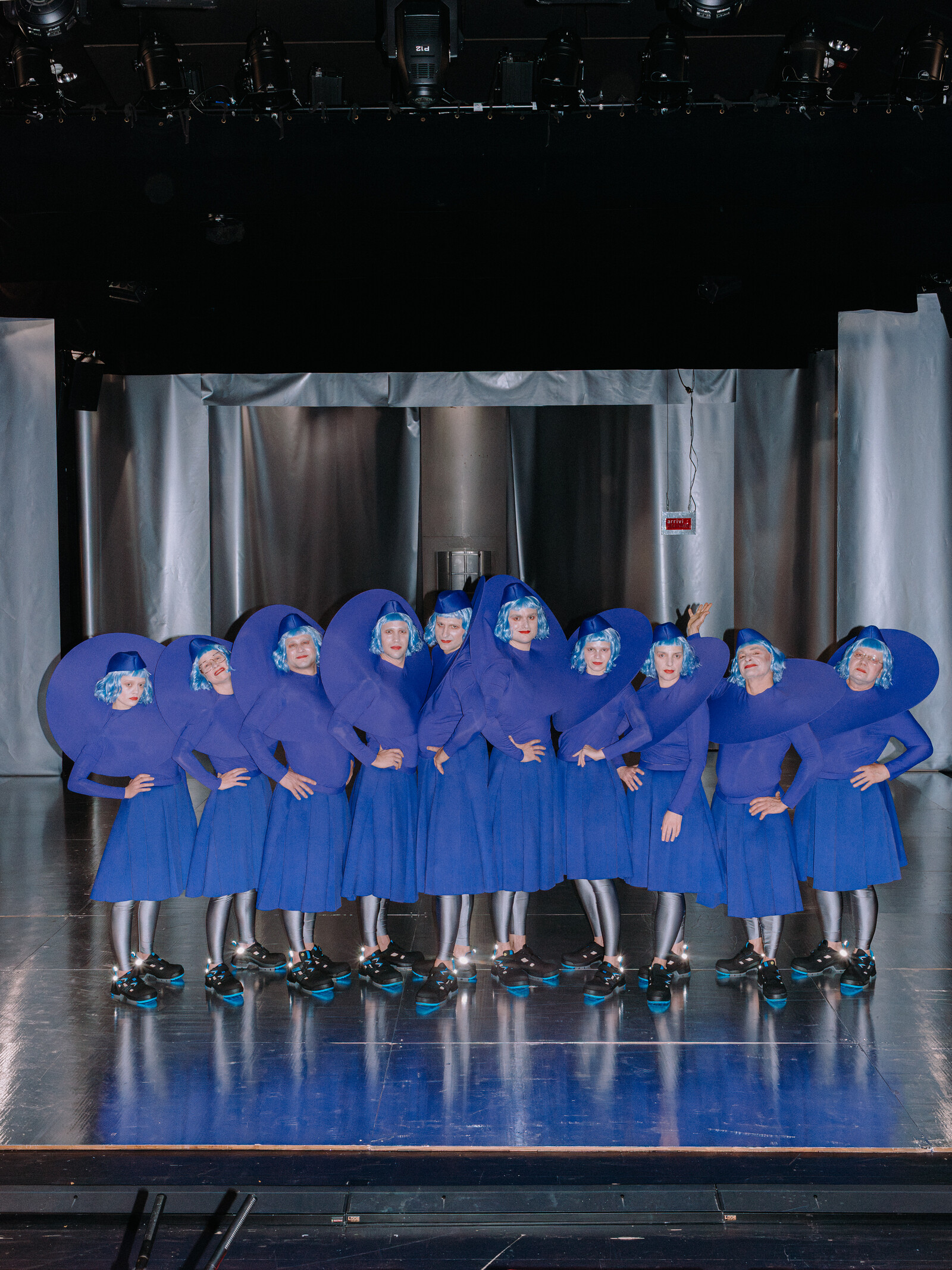 Das in blaue Kostüme gekleidete Ensemble posiert als Reihe aufgestellt im silbernen Bühnenbild.
