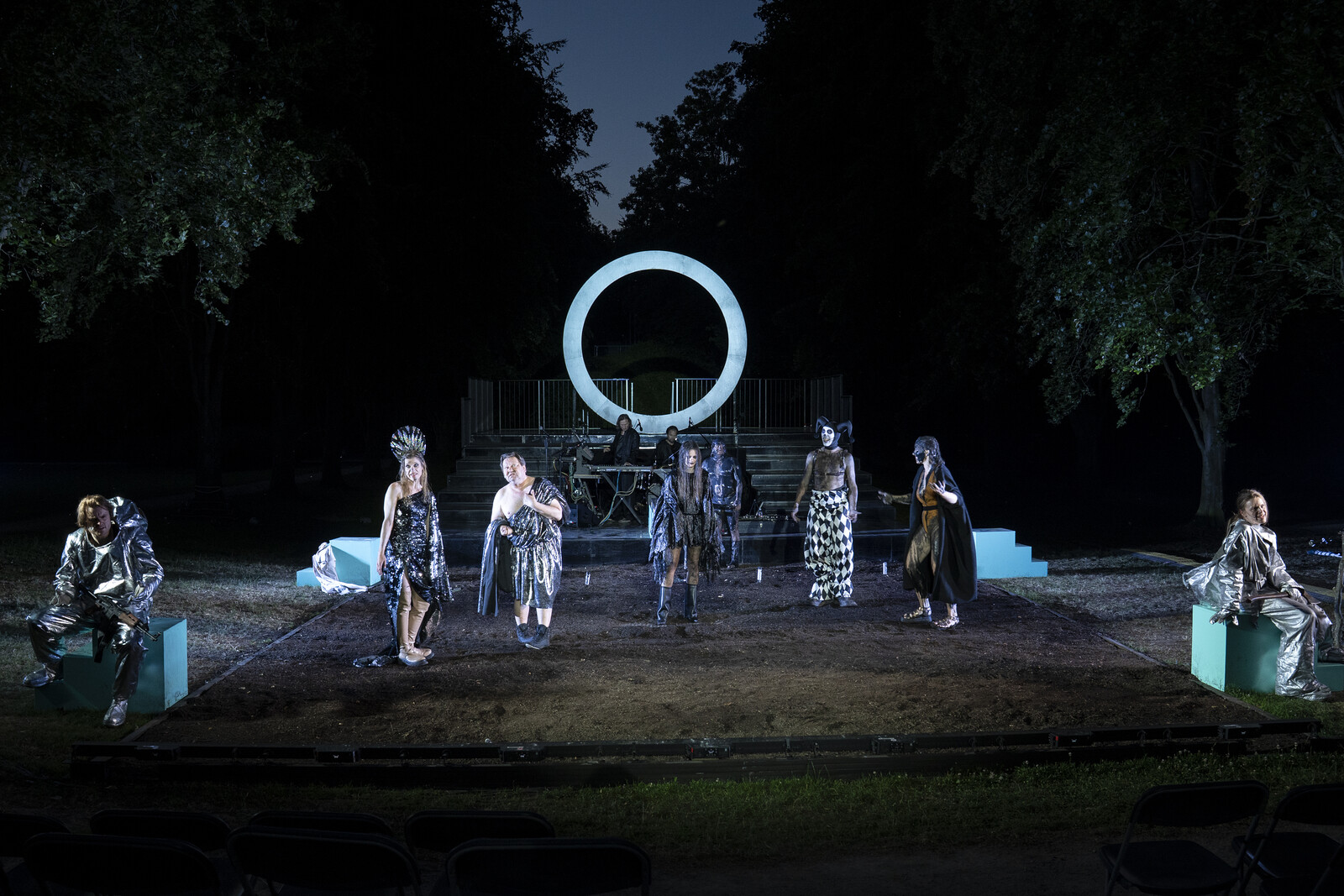 The open-air stage in Raffelberg Park for Circus Orestie, featuring all the actors and musicians, with a large, brightly lit ring in the background.