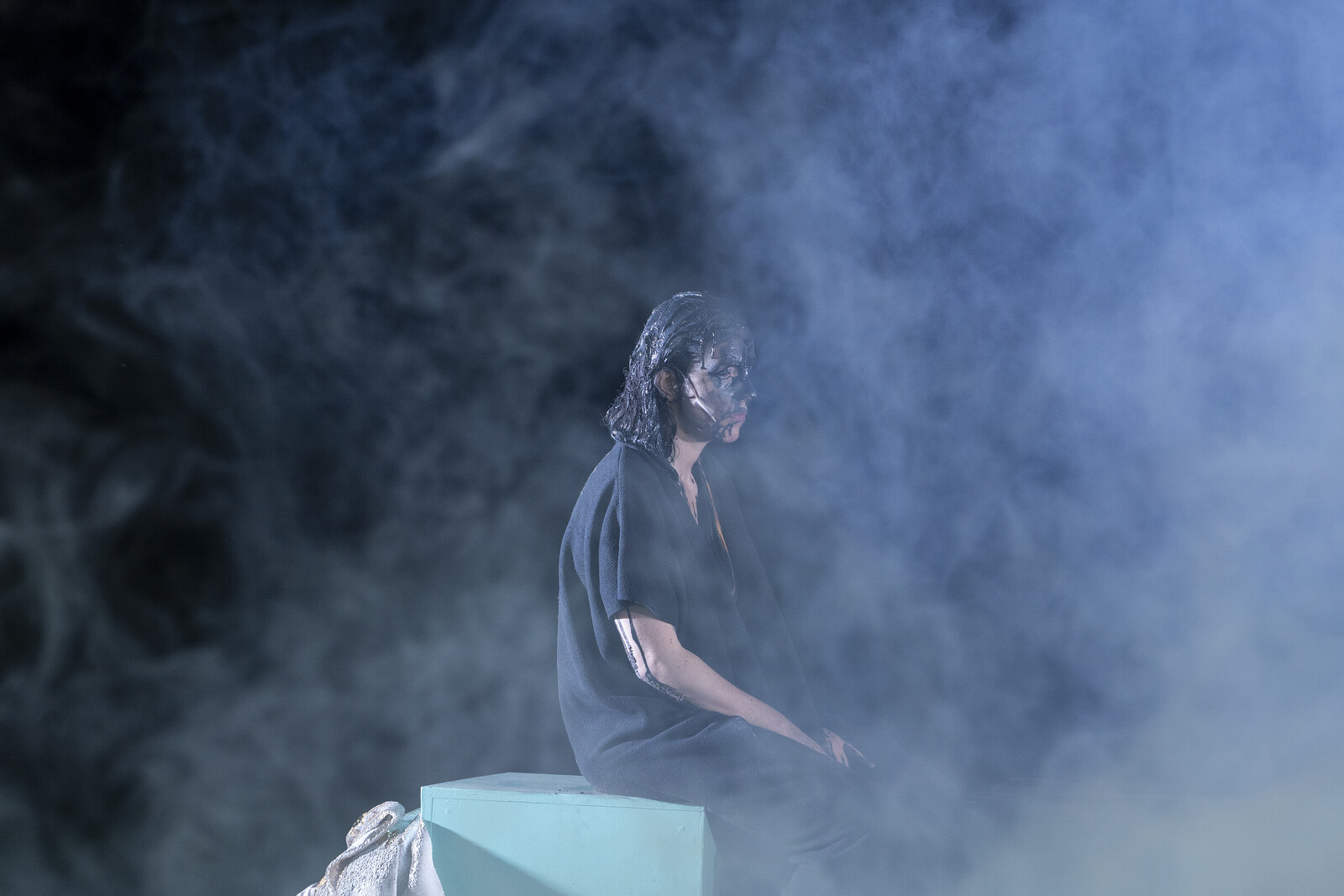 Kassandra (K. Schröder) sits on a platform in the mist, covered in oil.