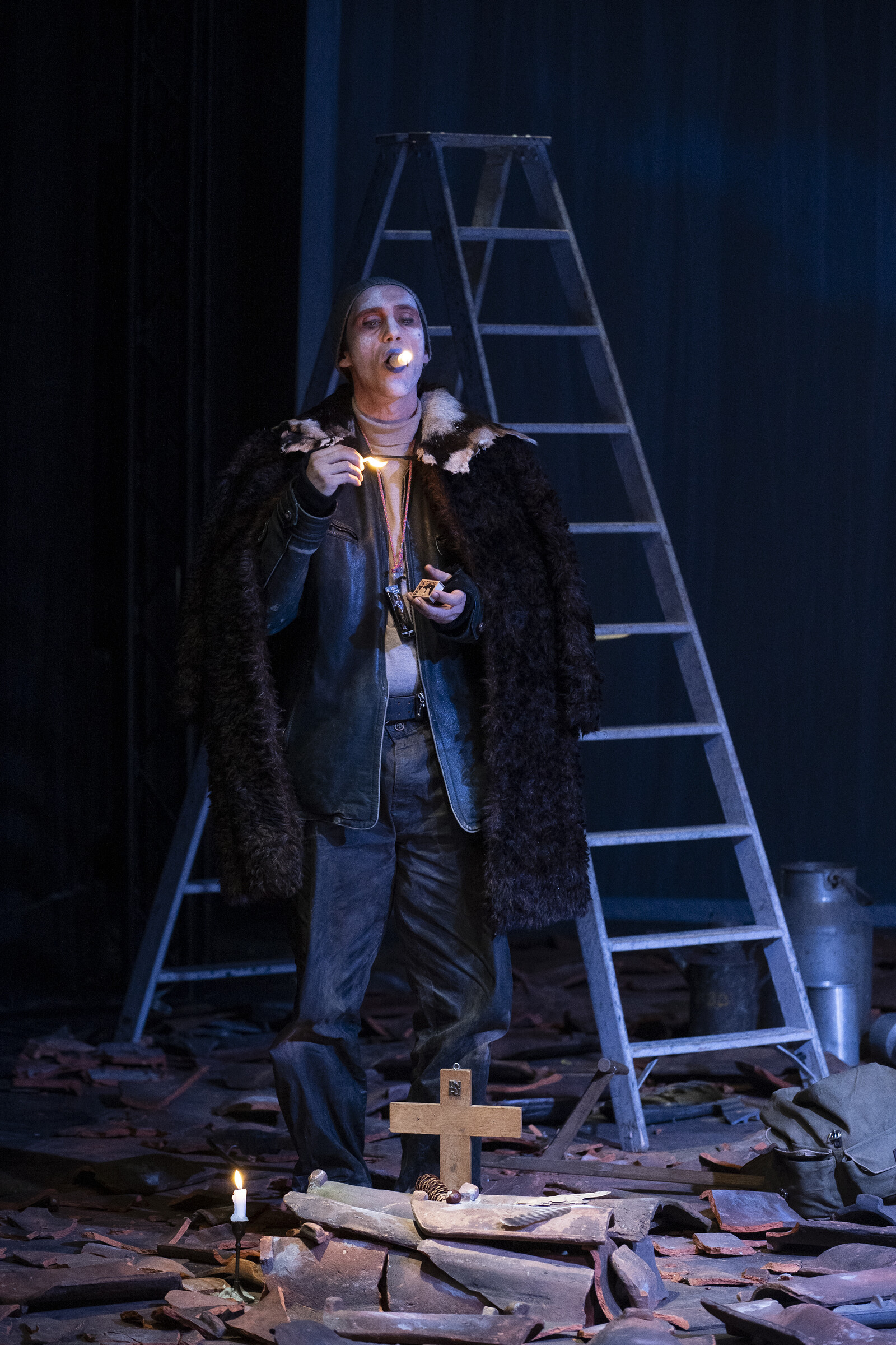 A man is standing in front of a ladder, holding a candle in his hand and with another in his mouth 