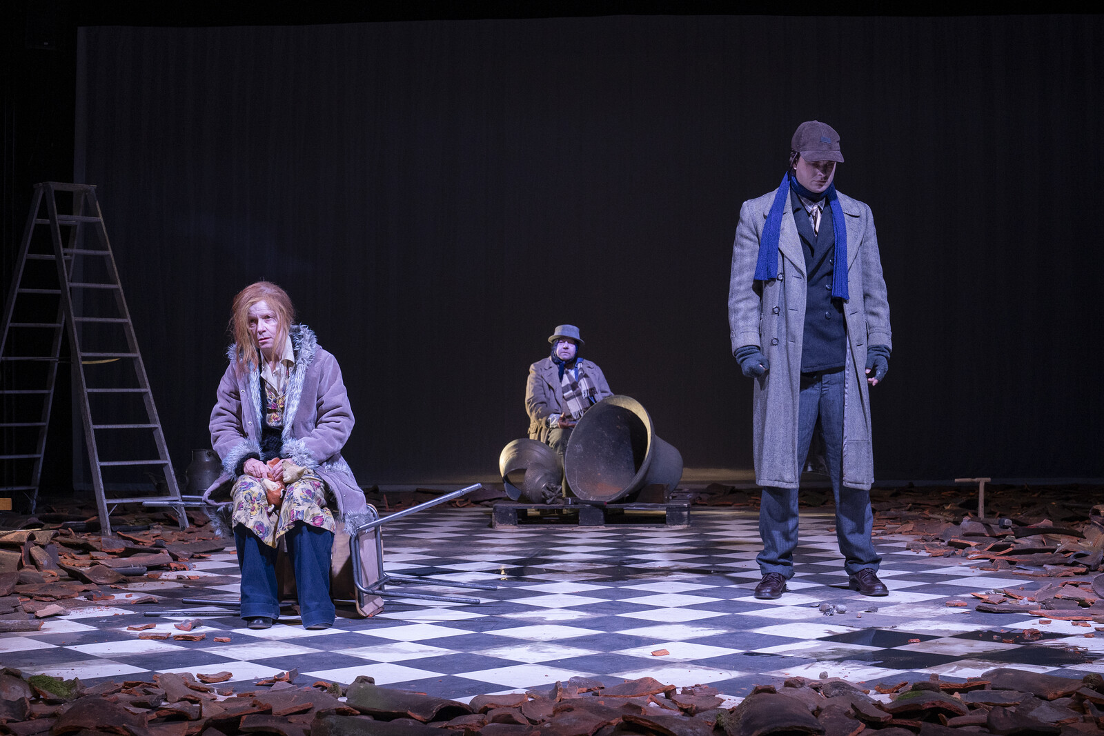 There are three people on stage: one is sitting at the back behind a church bell and two are standing at the front. 