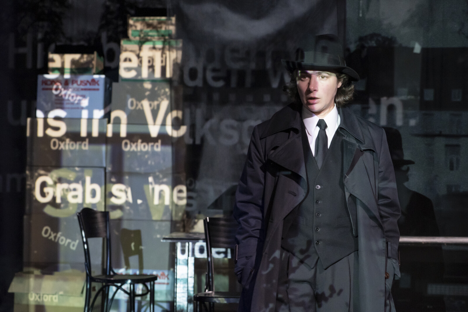 Eine als Mann gekleidete Frau steht vor Projektionen von Text an der Wand.