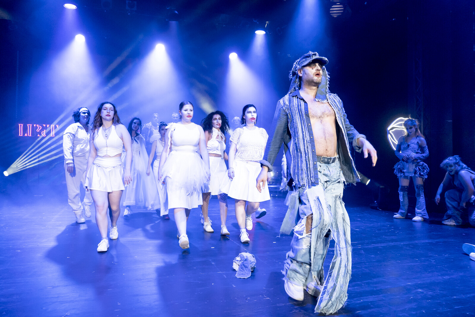 Ein Schauspieler in Jeanskostüm geht einer Gruppe weißgekleideter Statisten voran.