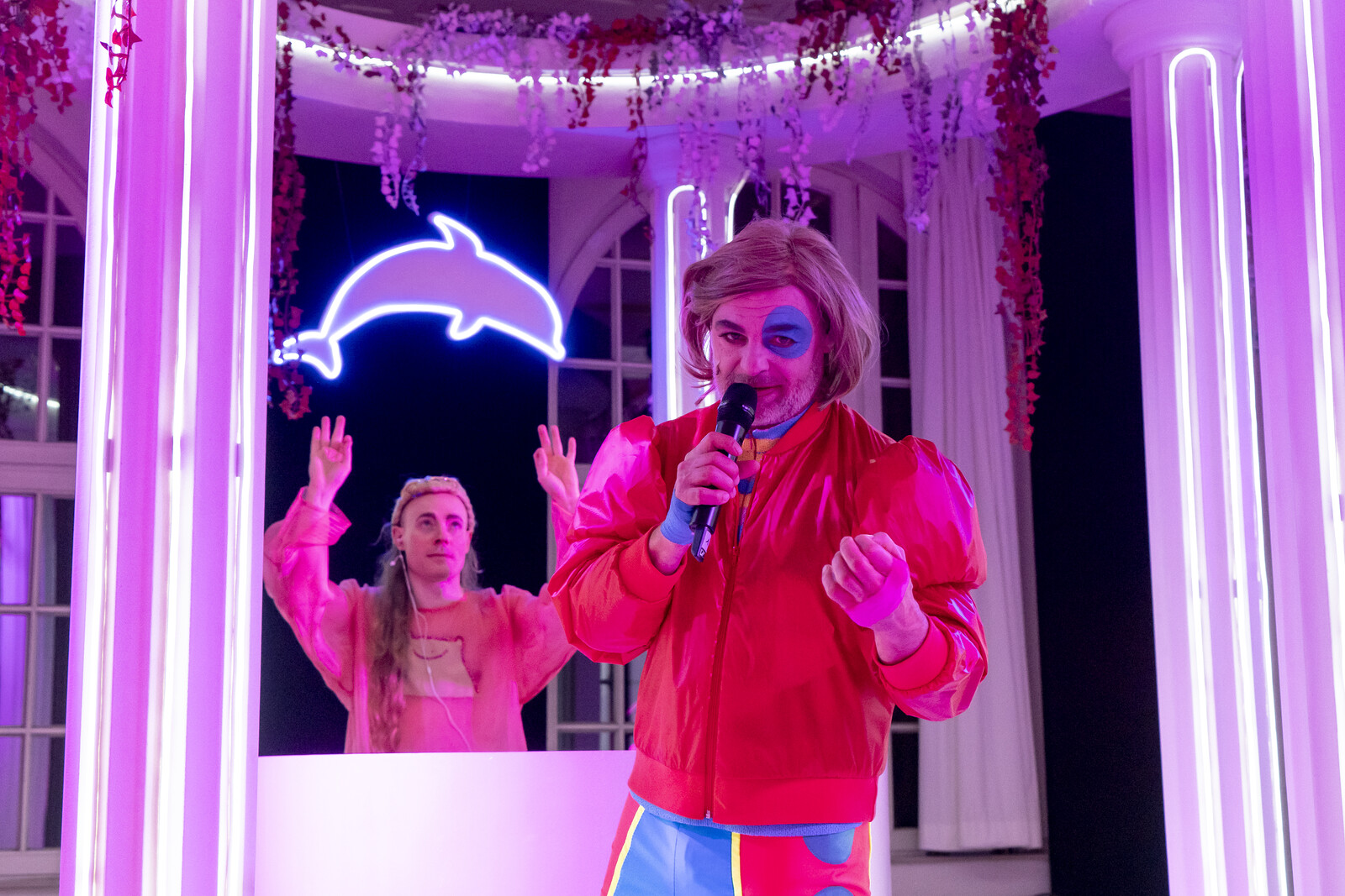 Der DJ steht hinter einem Pult in einer tempelähnlichen Struktur, davor ein Darsteller mit roter Jacke und Perücke mit Mikrofon.