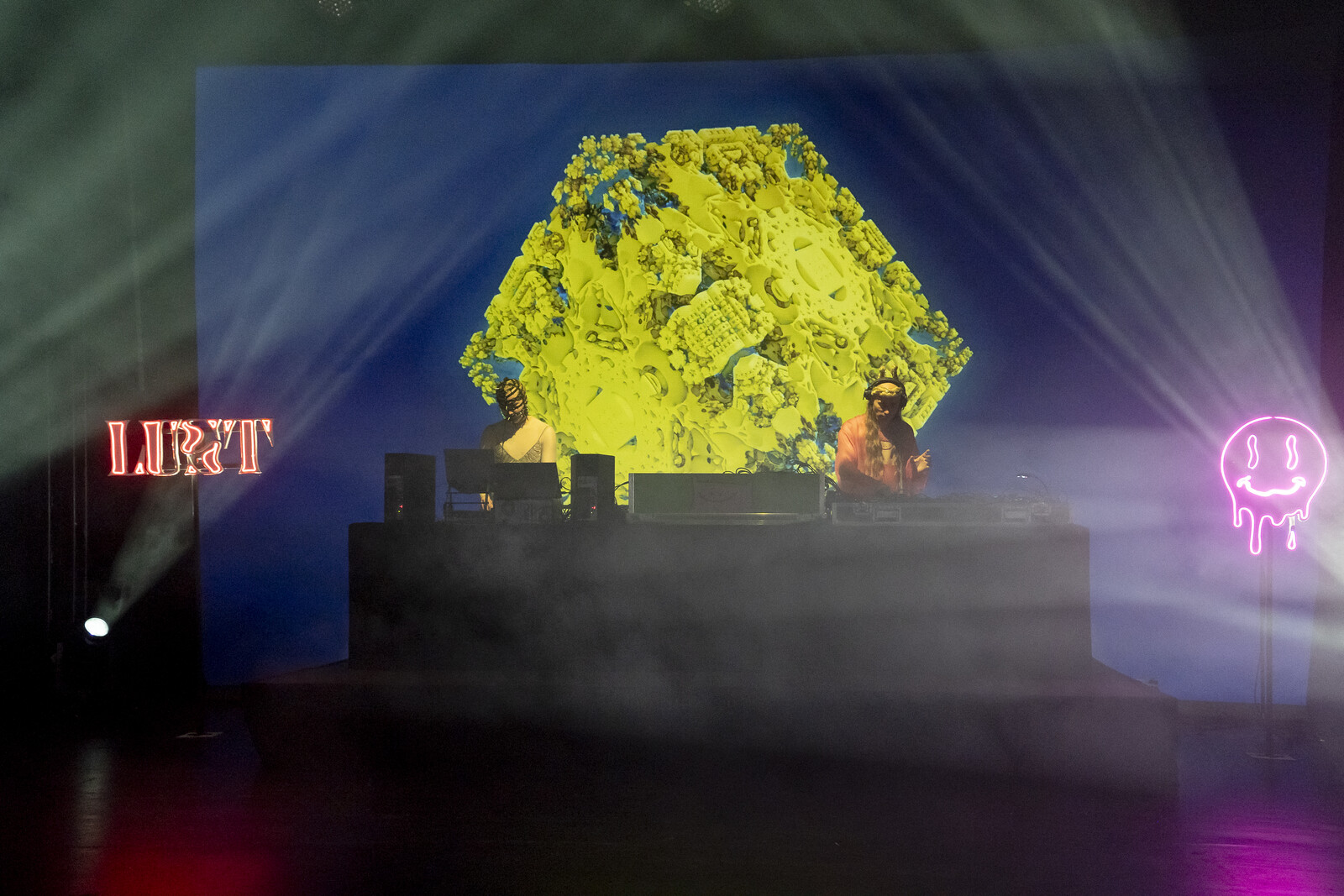 Ein dunkler Partyraum mit Tanzer*innen, Leuchtzeichen und DJ-Pult im Hintergrund.