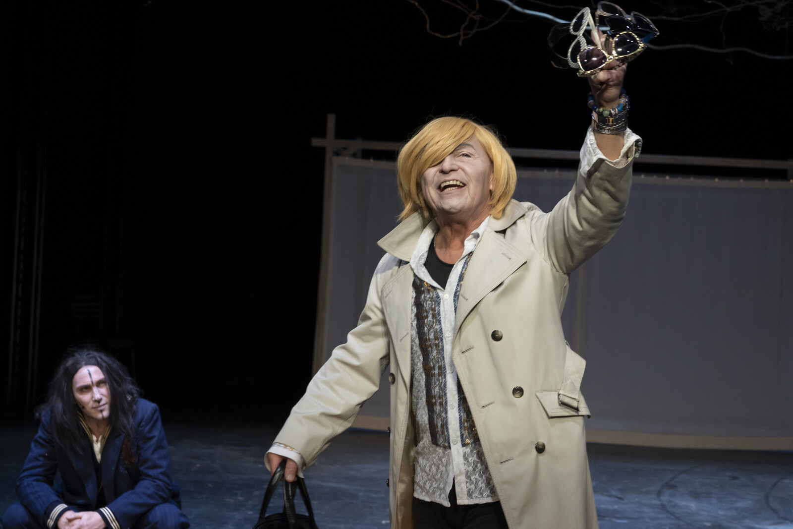 Ein Mann mit gelber Perücke preist Sonnenbrillen an und wird dabei von einem hockenden Mann beobachtet.