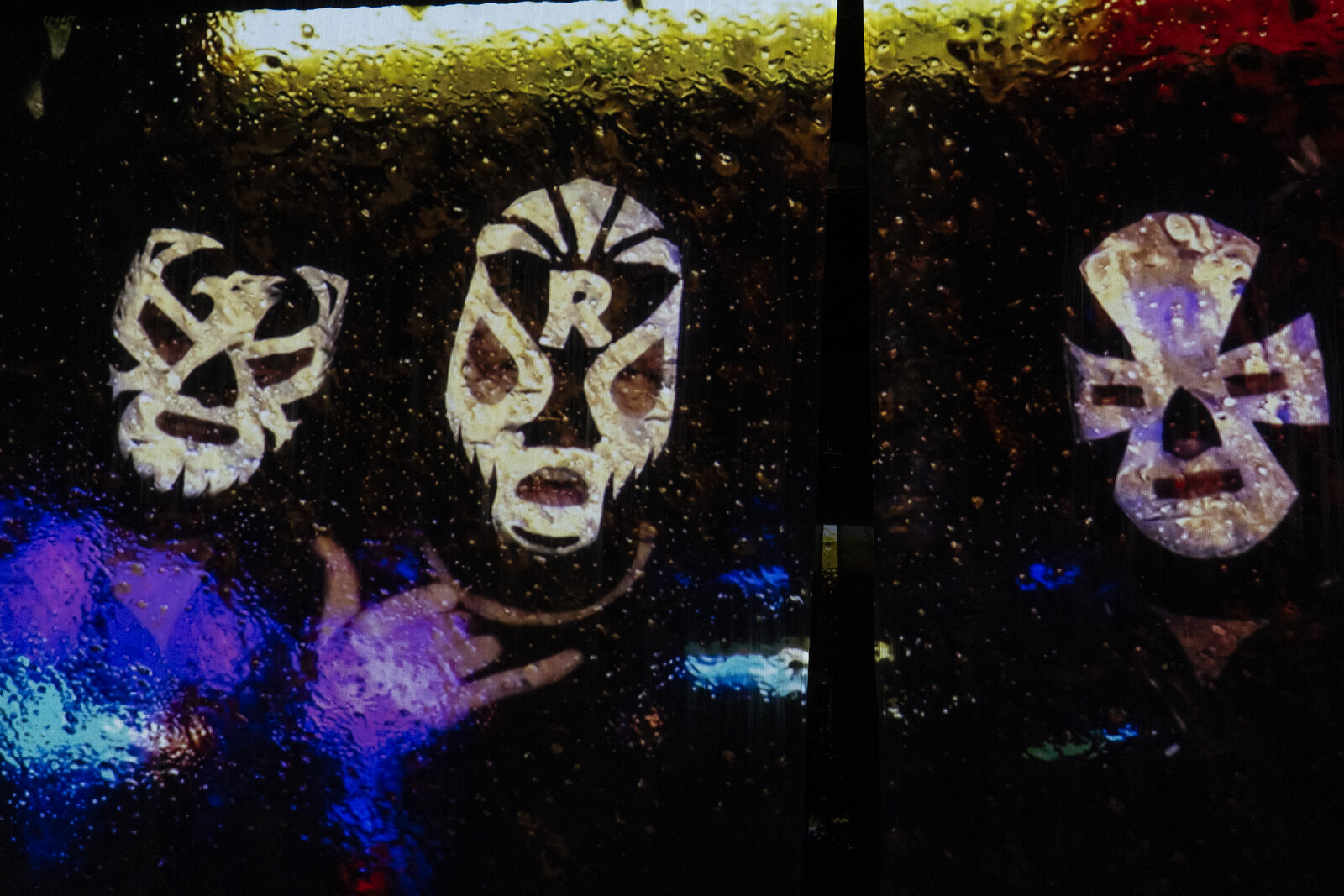 Auf einer verwaschenen Videoprojektion sind drei mit Wrestling-Masken verhüllte große Köpfe zu sehen.