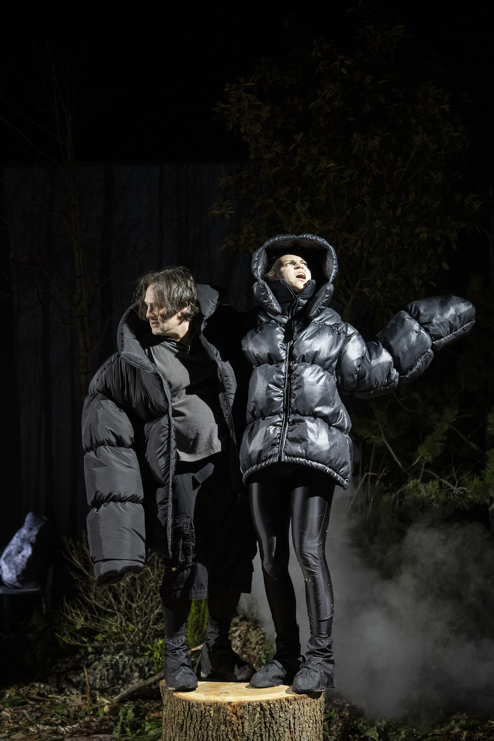 Zwei Darsteller stehen in überdimensionierten Daunenjacken auf einem Baumstumpf.