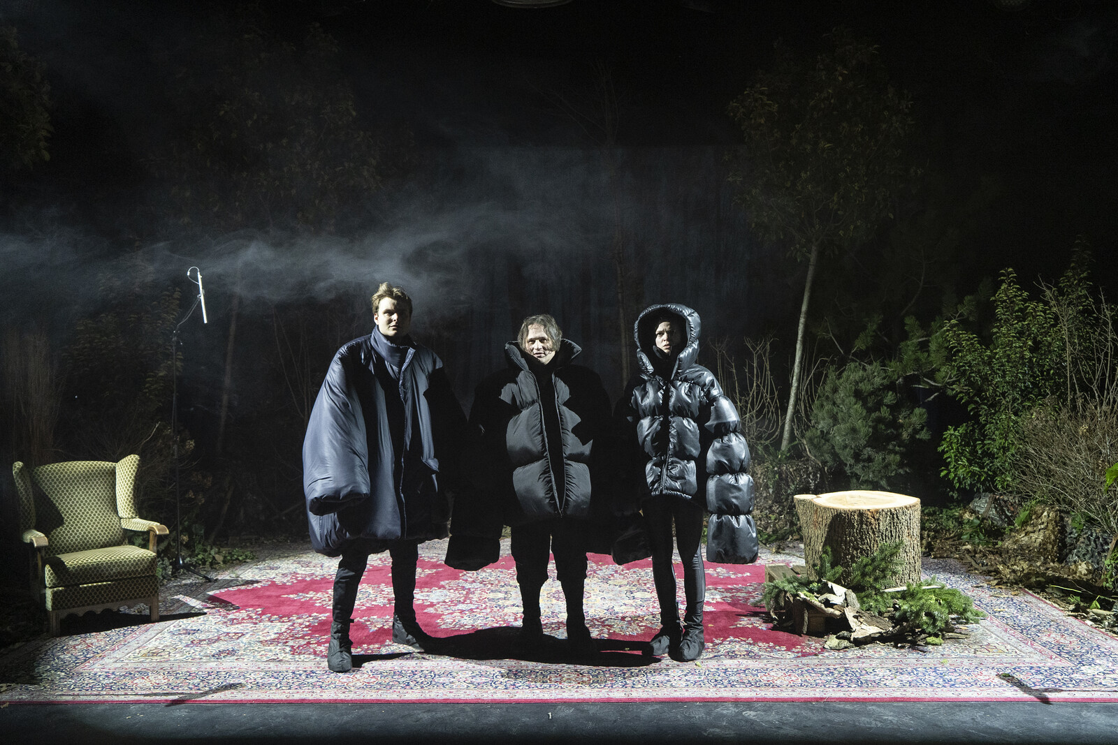 Drei Darsteller in überdimensionierten Daunenjacken stehen auf einem Perserteppich, umgeben von Bäumen.