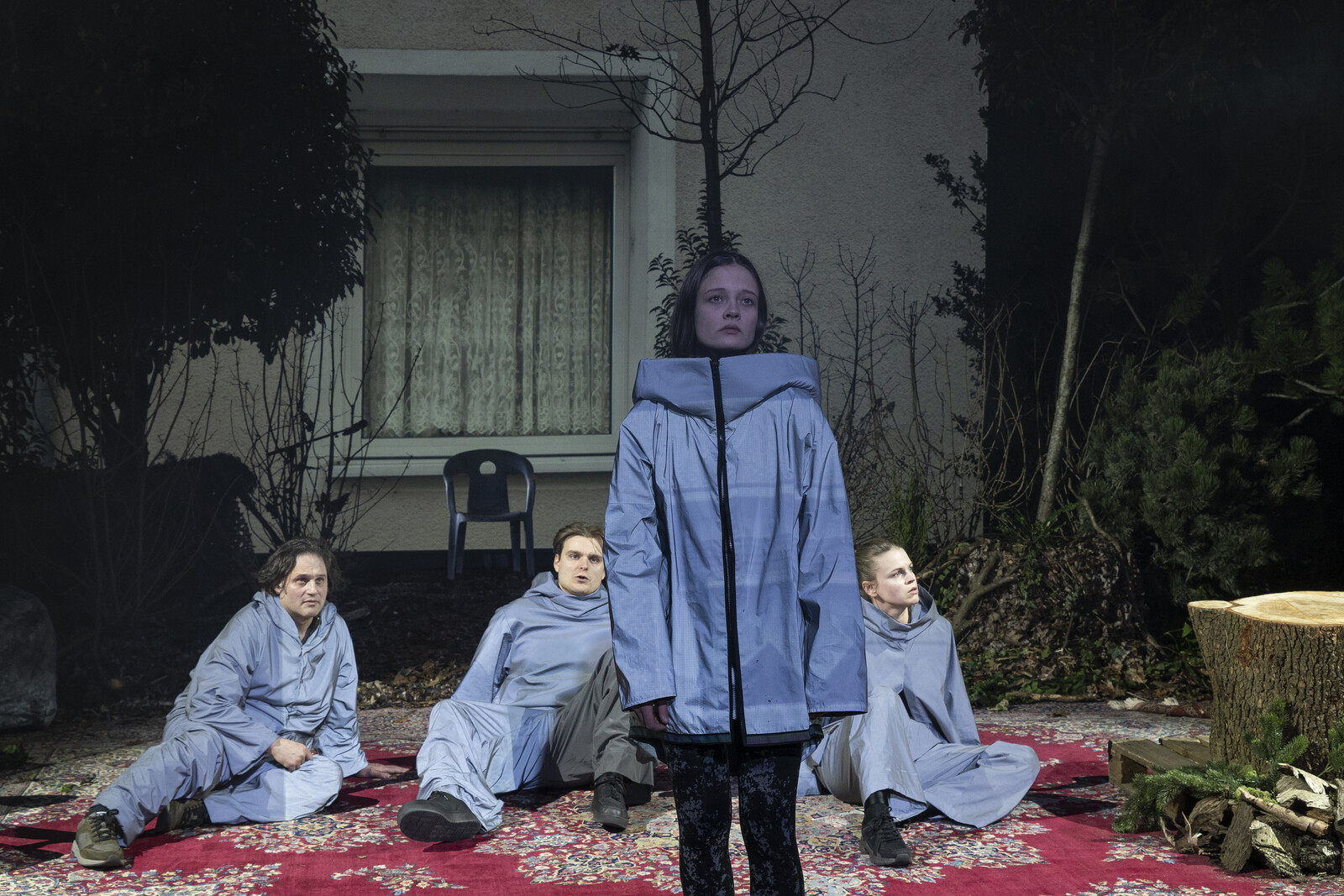 Drei Darsteller in Trainingsanzügen liegen auf einem Perserteppich, vor ihnen steht ihnen abgewendet im Vordergrund eine junge Frau in gleicher Kleidung.