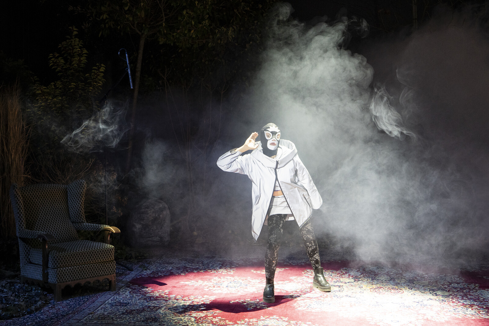 Eine Gestalt mit einer Wrestling-Maske und in reflektierender Kleidung steht in der Bühnenmitte und macht eine Geste des Lauschens.