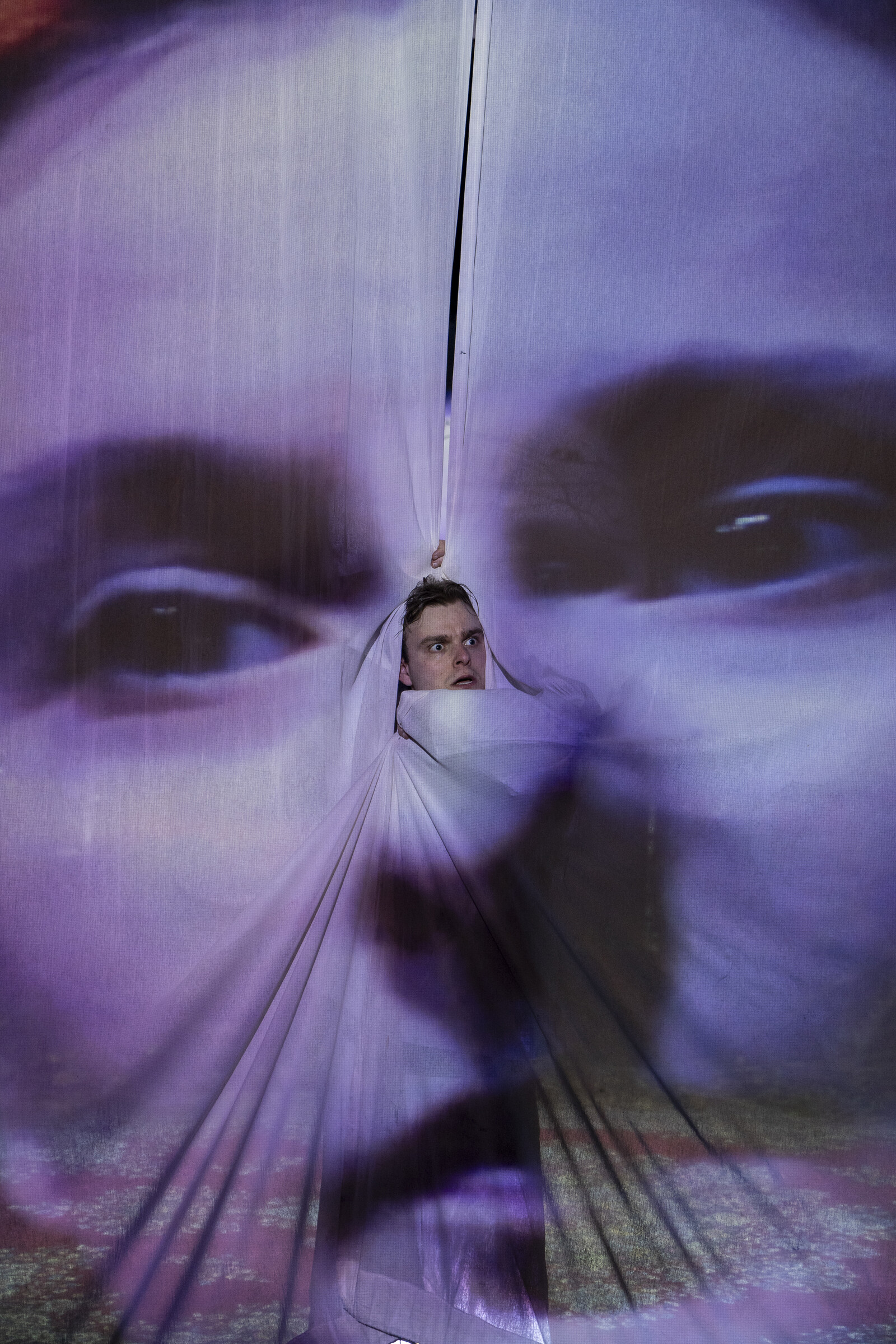 Ein Schauspieler schaut aus einem Vorhang hervor, auf dem ein riesiges Gesicht einer Frau projiziert ist.