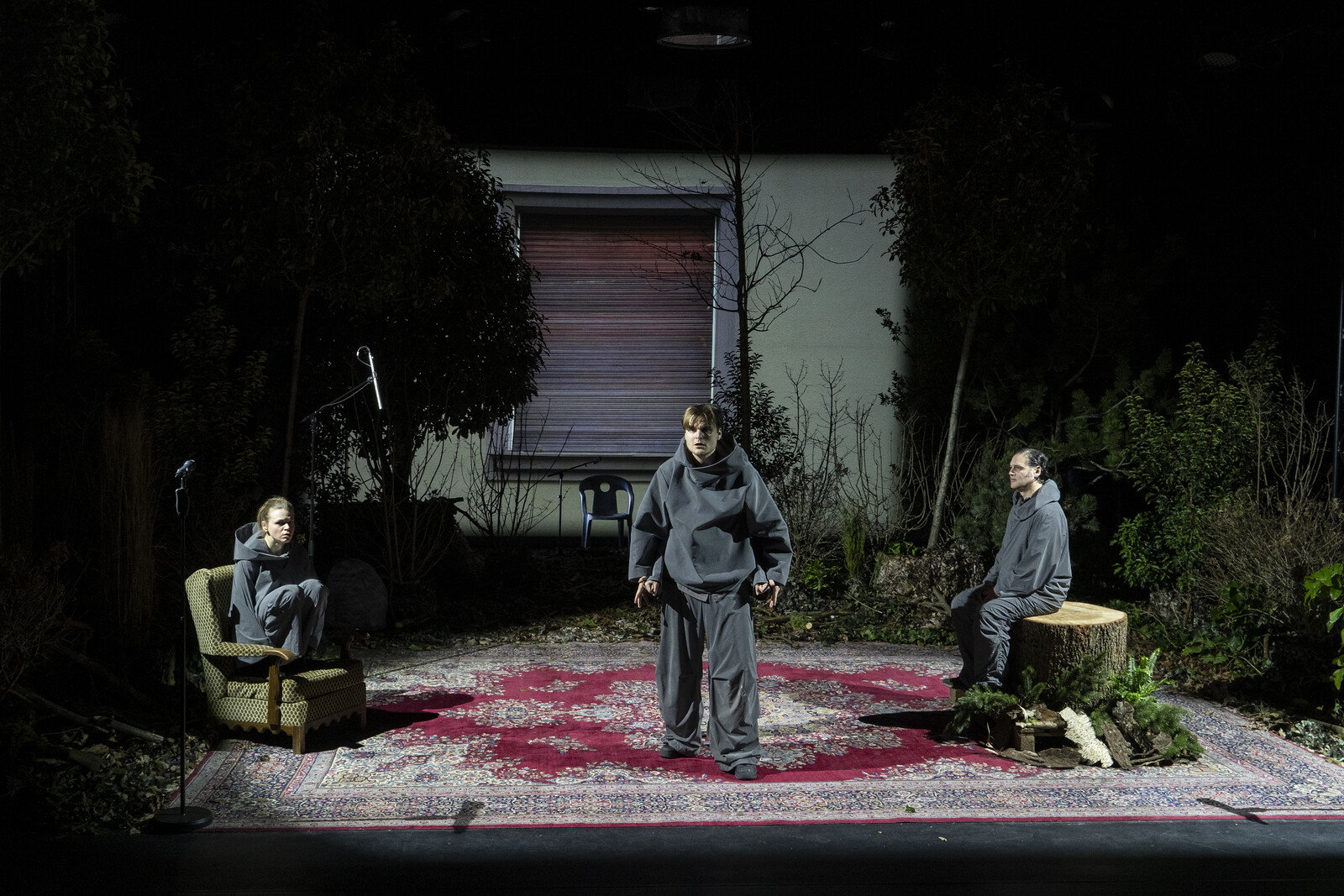 Drei Darsteller in grauen Trainingsanzügen stehen auf einem großen Perserteppich, von Bäumen umgeben, vor der Projektion einer Hausfassade.
