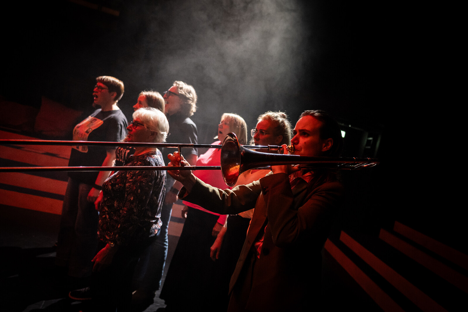 In the foreground, a woman with a trumpet; in the background, a citizens’ choir of six people stands.