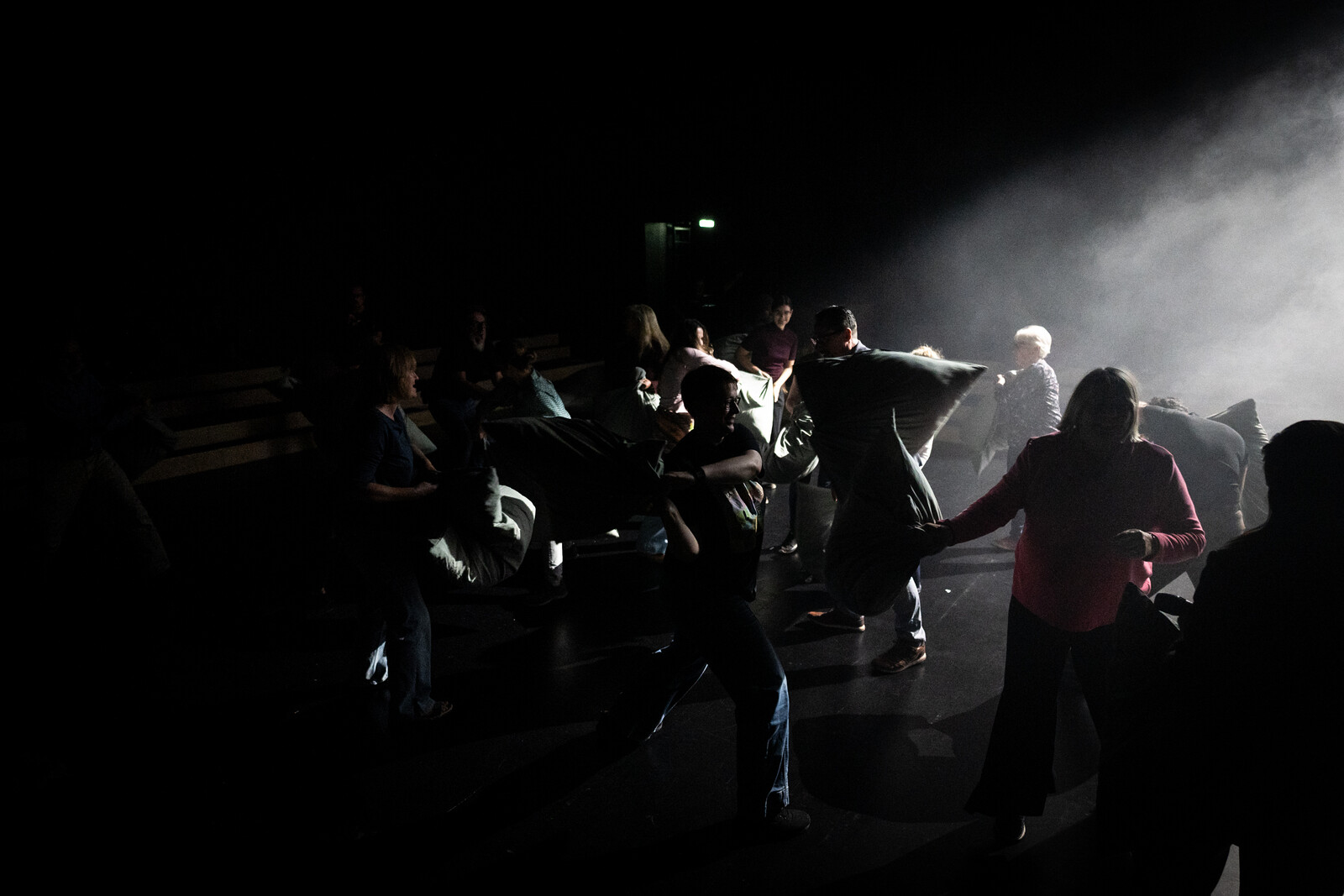 A group of people on a dark stage engage in a pillow fight, surrounded by theatrical haze.