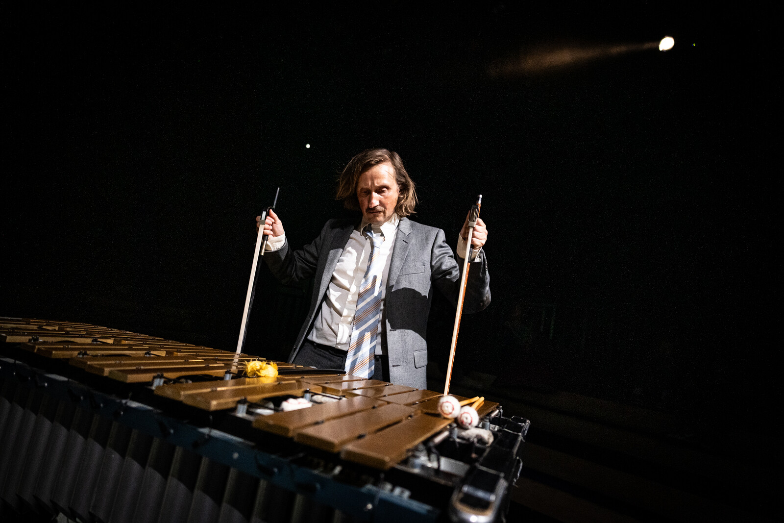 A man in a suit plays a vibraphone with a focused expression, holding two violin bows in his hands.