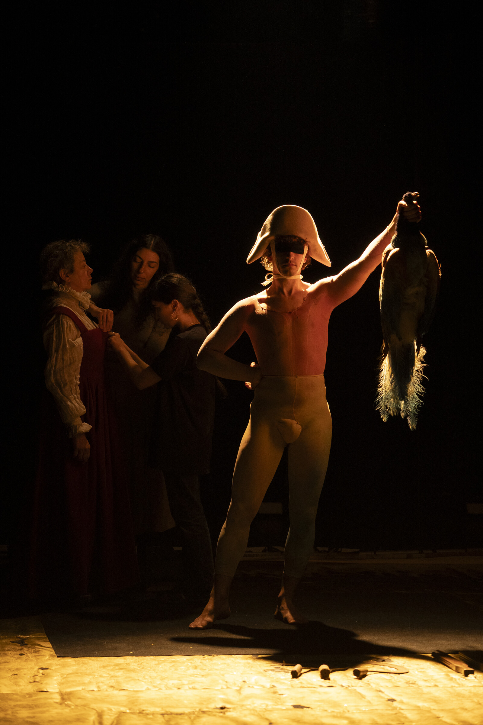 A man in a jester's cap holds up a dead bird, behind him three women in the dark background.