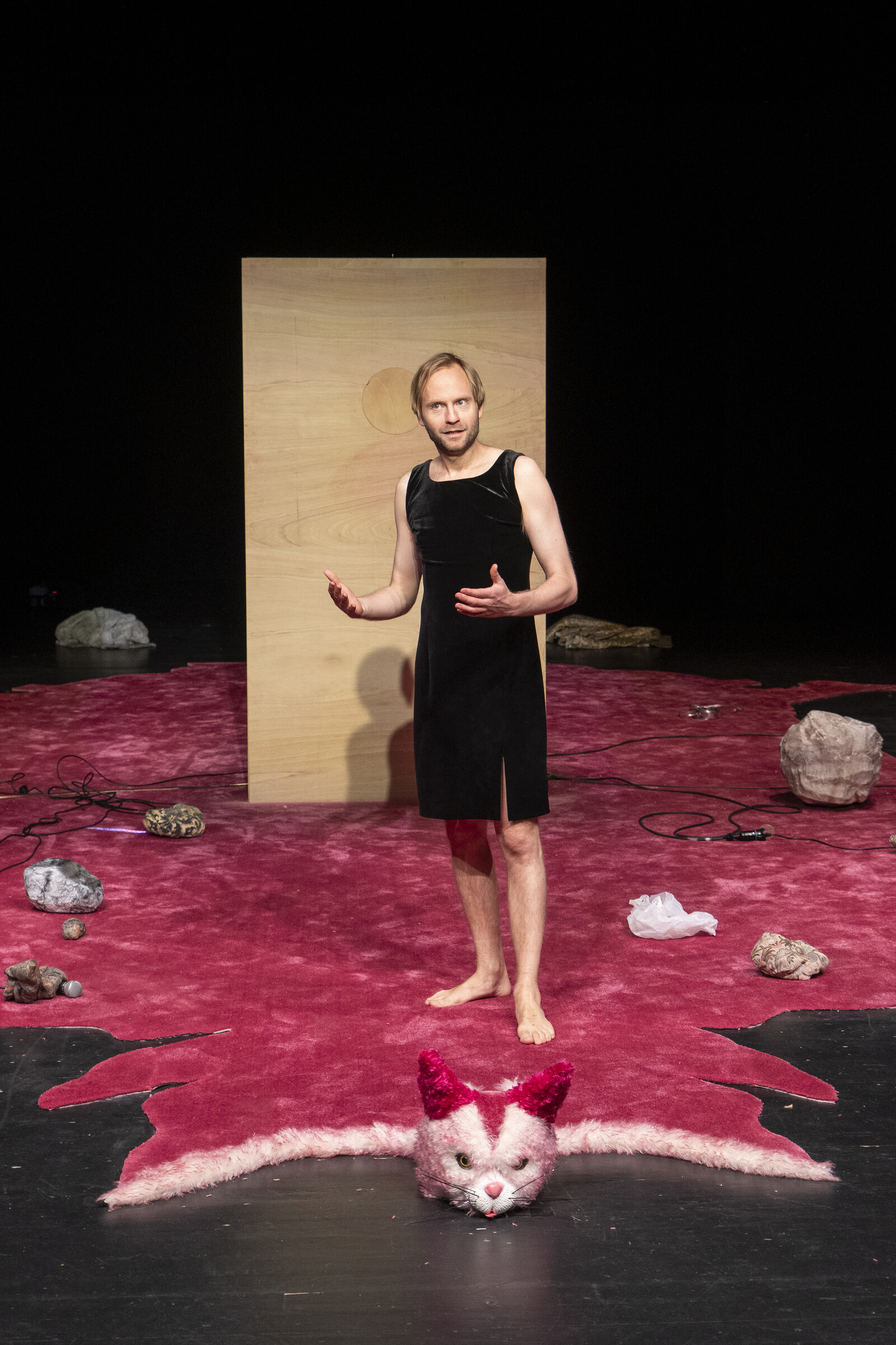 An actor in a black dress stands on a pink carpet in front of a large wooden box.