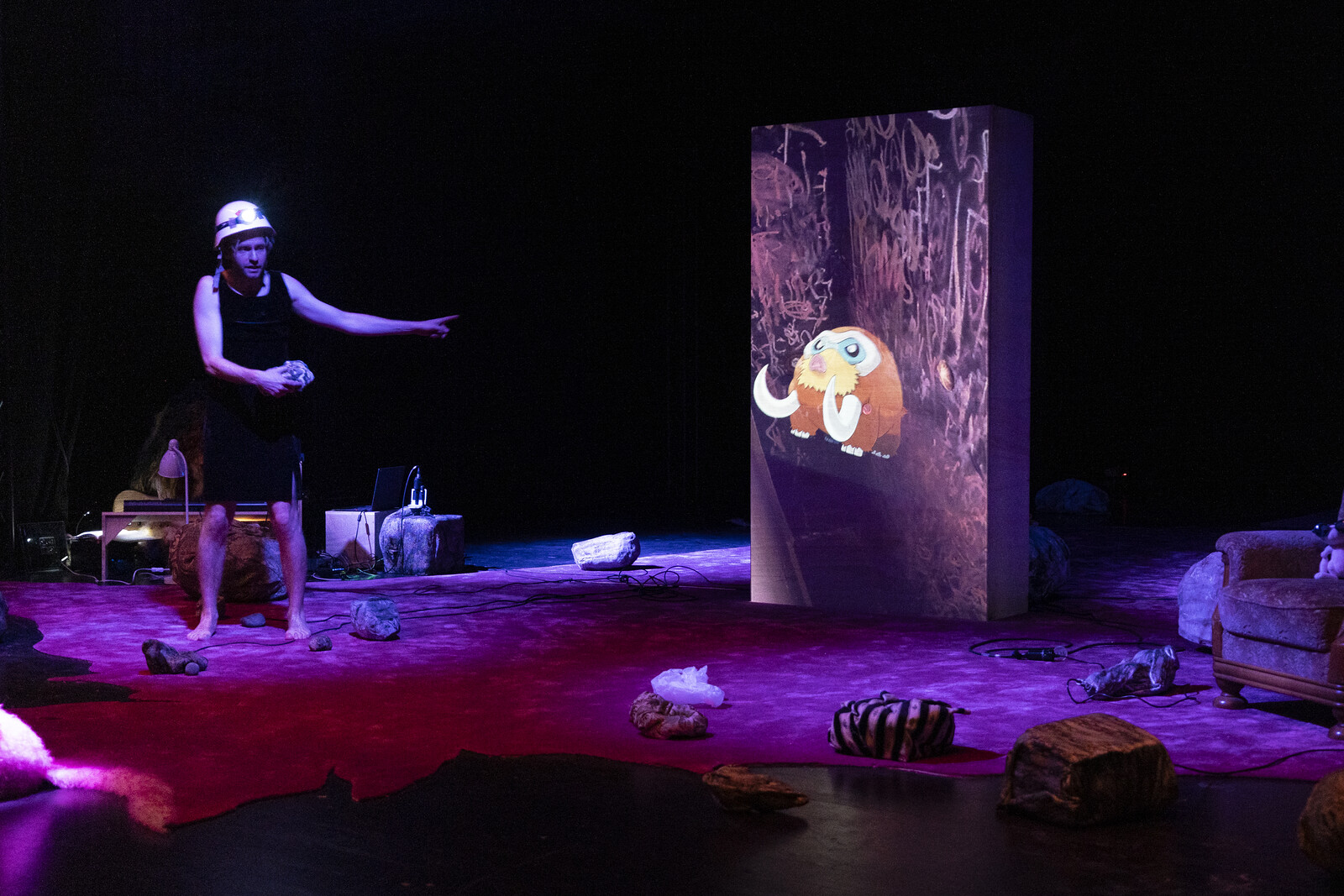 An actor wearing a dress and a protective helmet projects the image of a small mammoth onto a large wooden box.