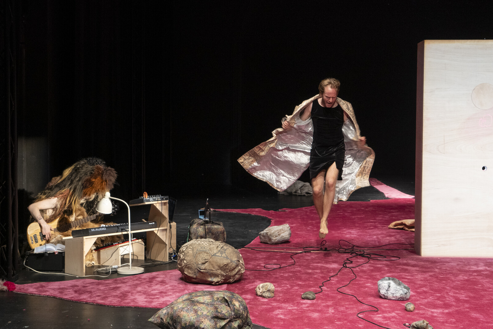 The performer, dressed in a dress and coat, jumps on a pink carpet, while next to him a musician sits in a huge fur costume and plays the guitar.