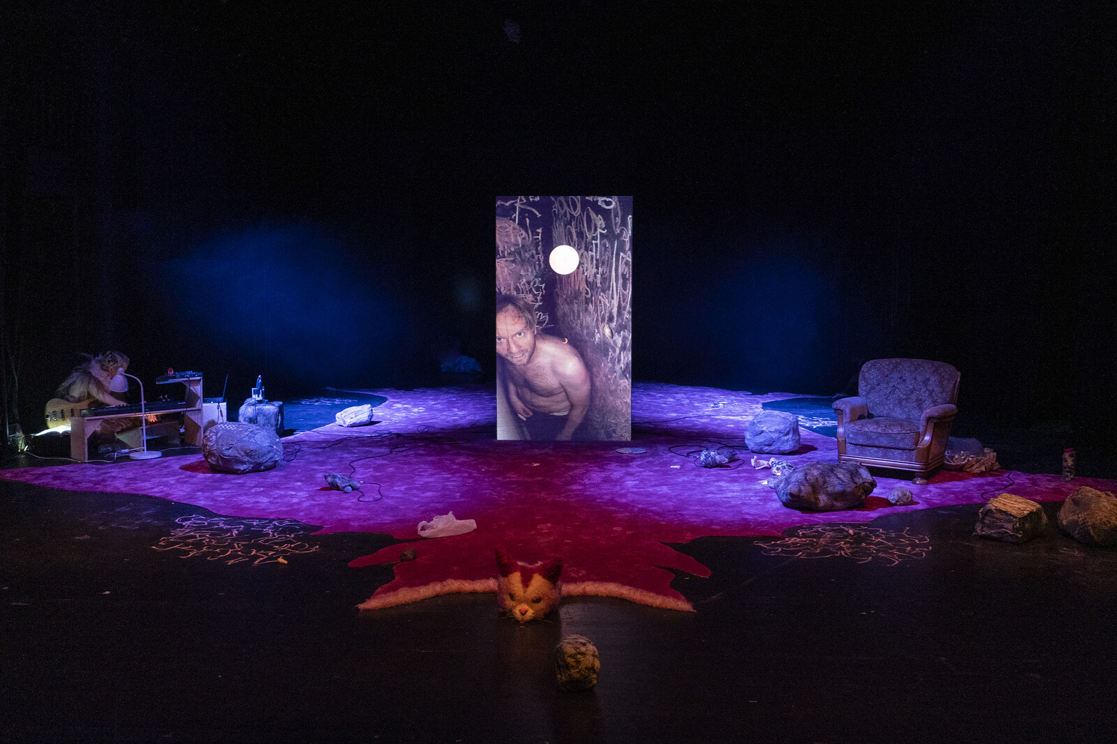 An image of the performer in a cave is projected onto a wooden box standing on a pink carpet.