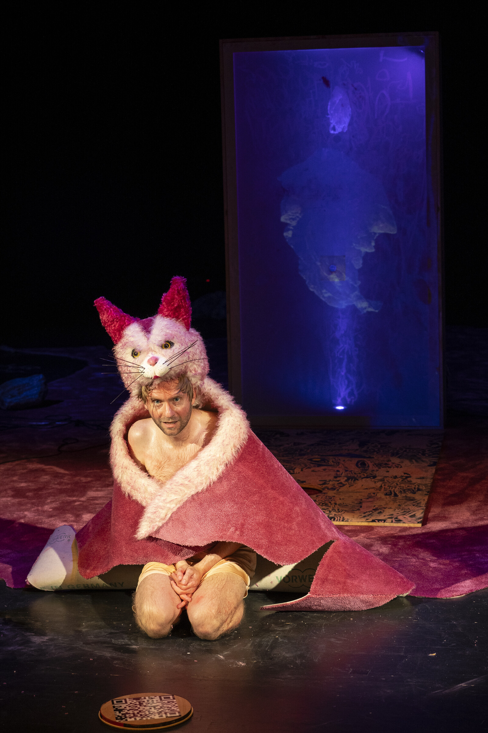 The actor is crouching on the floor, wrapped in a pink cat rug.