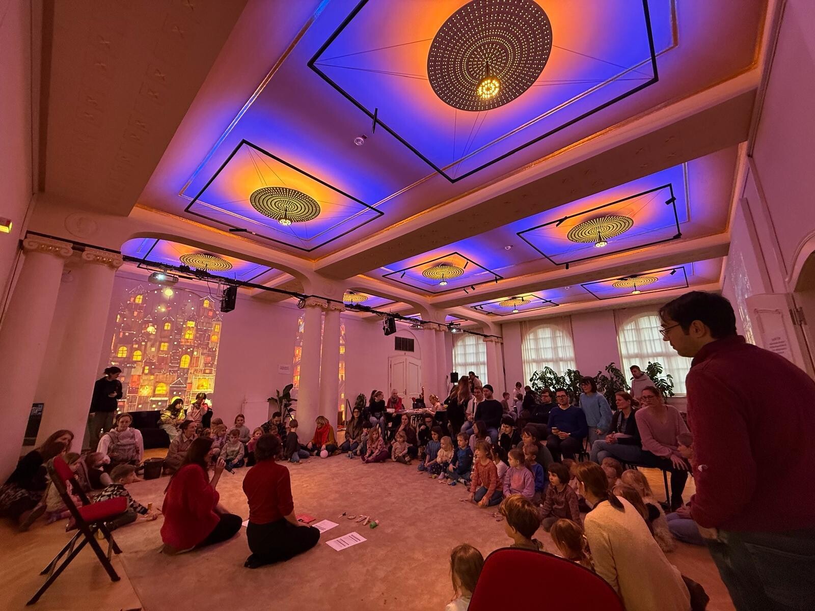Many children are sitting in the colorfully lit theater lobby; their parents are there too, listening to two people kneeling on the floor.