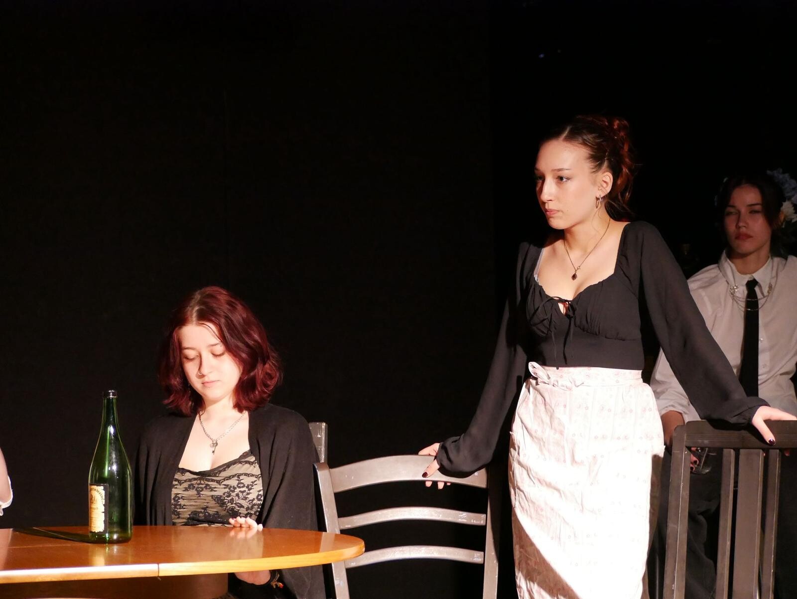 Two people are on stage; one is sitting at the table and one is standing at the table.