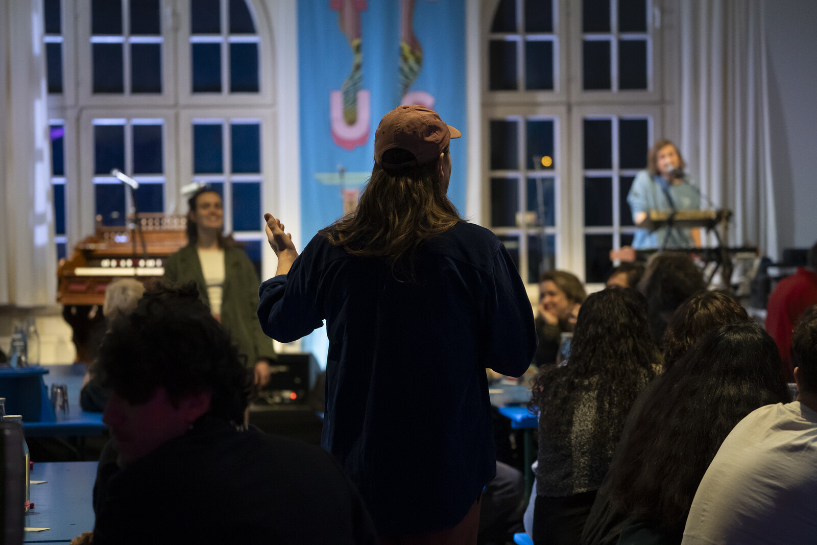 A performer stands in the audience with his back to the camera and looks at the stage with musicians.