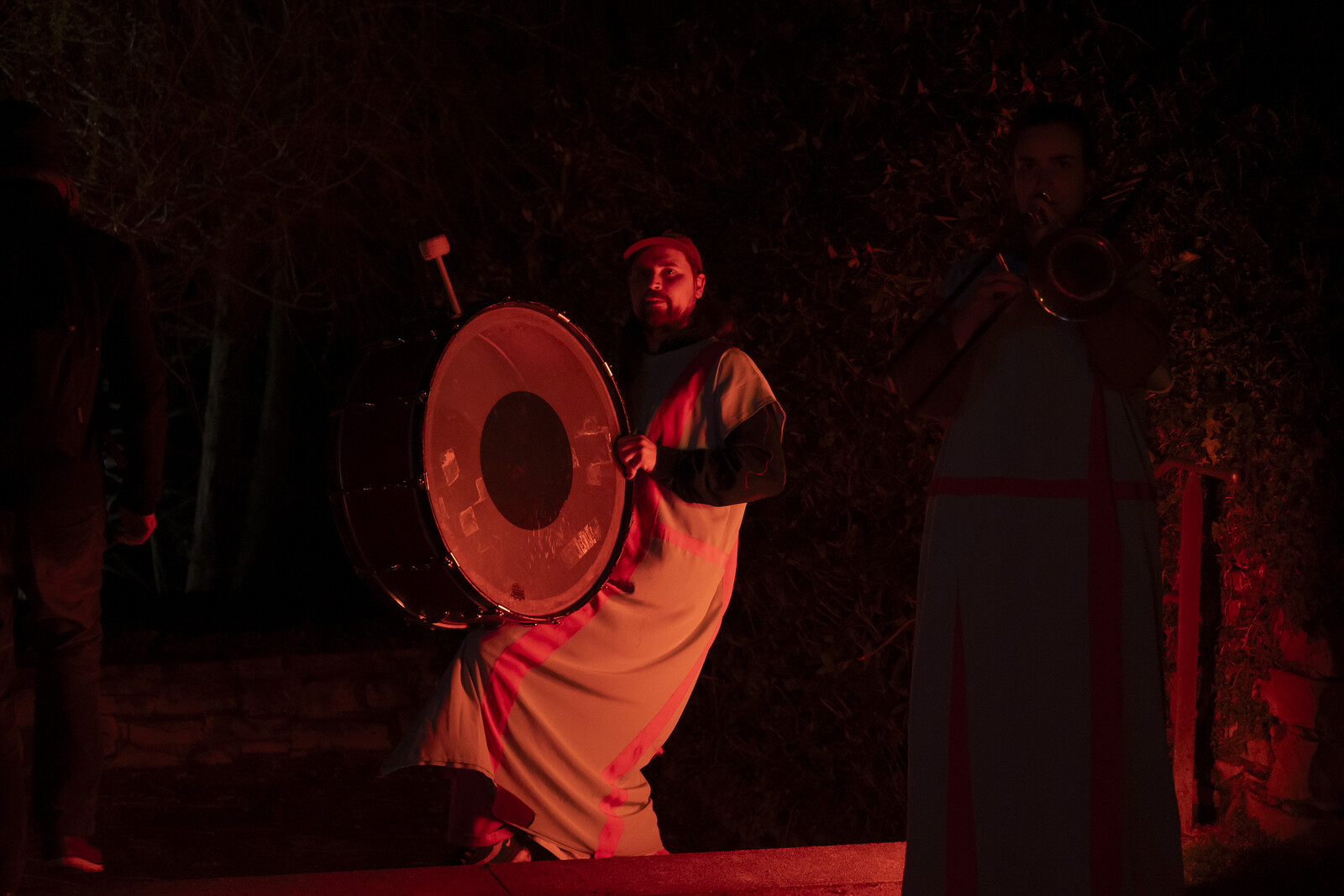 A performer in a frock stands in the semi-darkness and beats a large drum.
