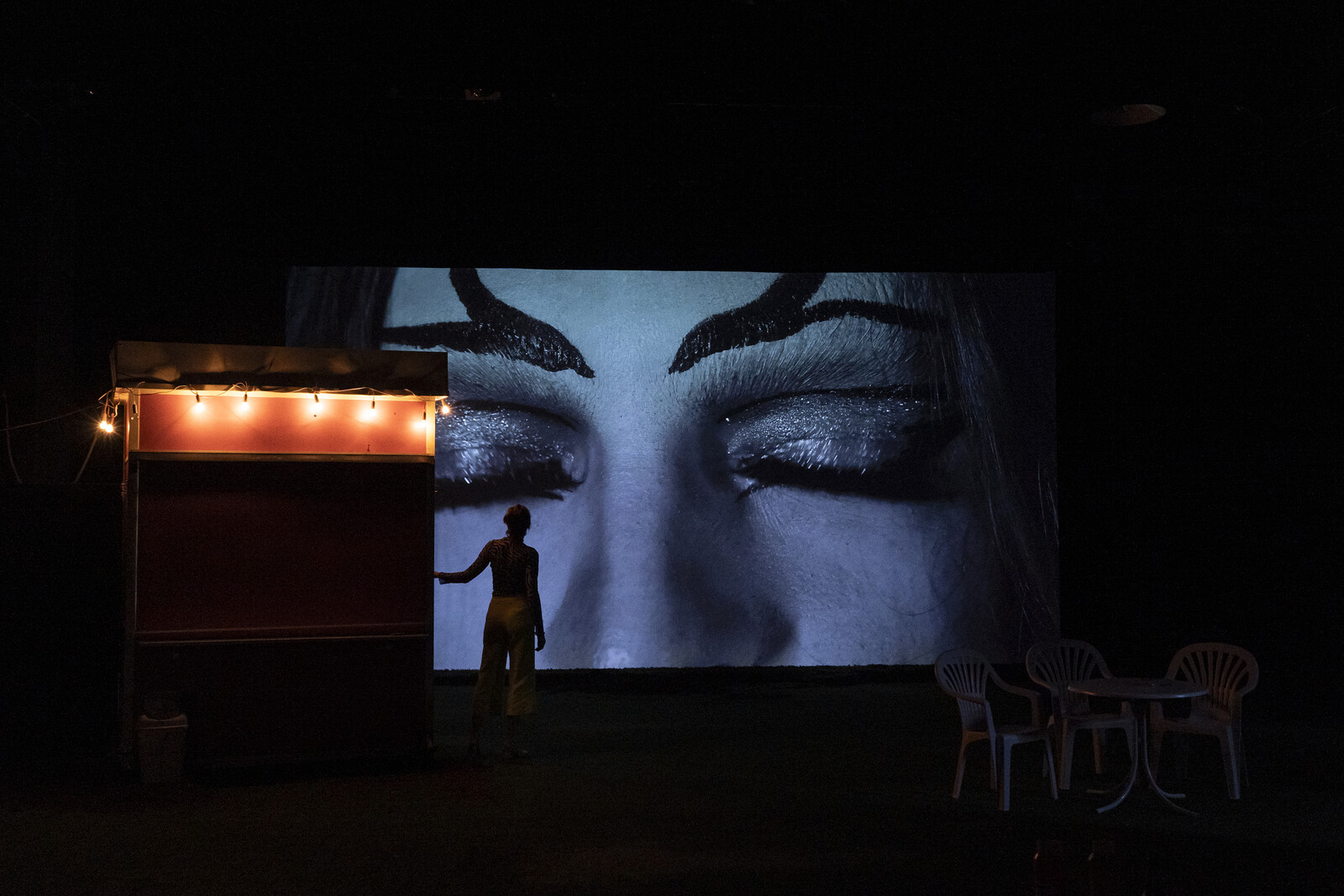 On a dark stage, a woman looks at a large projection in the background showing a pair of closed eyes.
